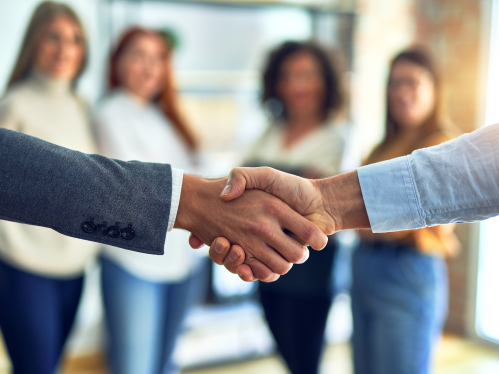 Group of business women standing together watching two colleagues shaking hands at the office