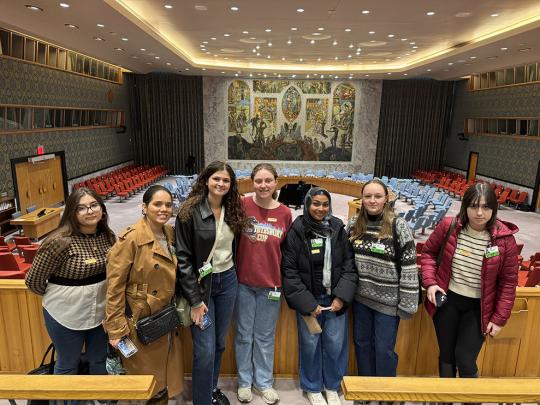 Students stand in large assembly room with art piece covering wall
